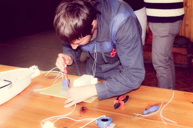 A high school student is meticulously soldering wires onto a circuit board of a small robot, with a focused expression. The workspace is organized with various electronic components, tools, and a technical diagram. The student is wearing safety glasses and demonstrating precision in their task.
